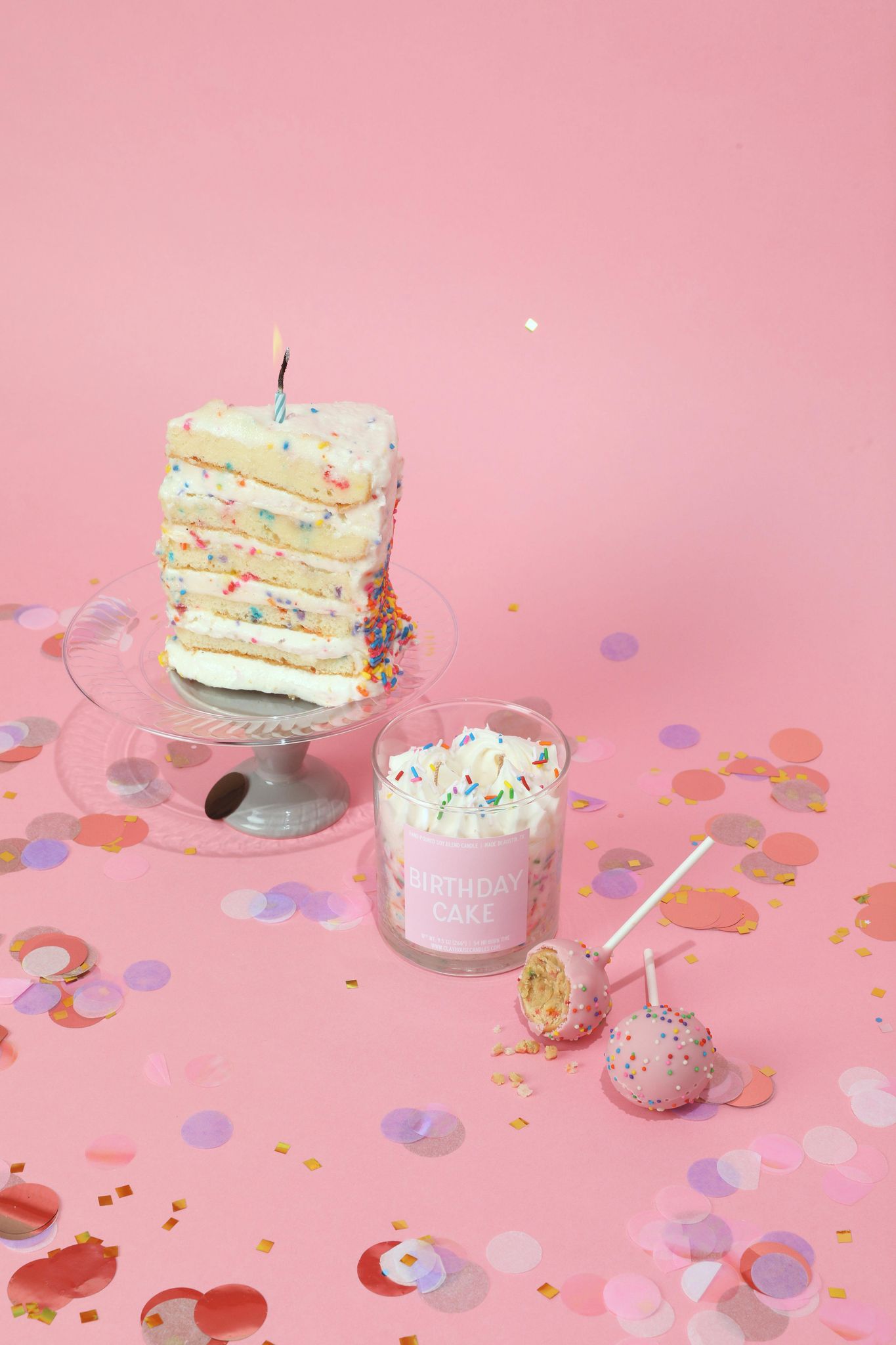 Layered cake with a candle, glass of milkshake, and lollipop on a pink background with confetti.