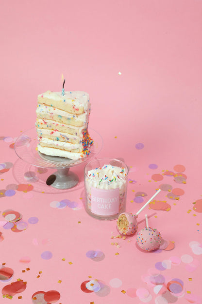 Layered cake with a candle, glass of milkshake, and lollipop on a pink background with confetti.