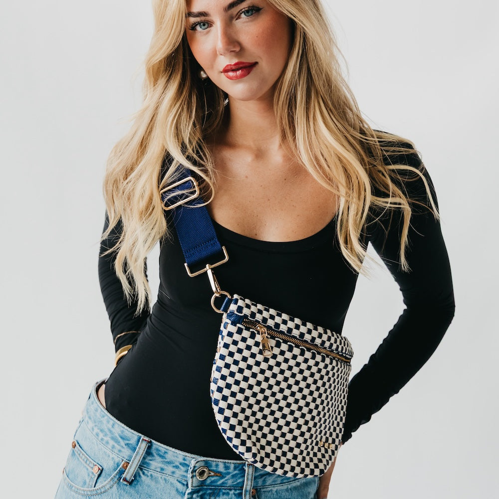 Woman wearing a black long-sleeve top and blue jeans with a checkered bag over her shoulder on a light gray background