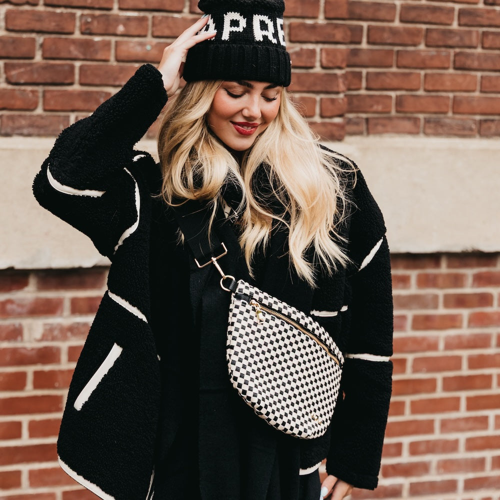 Woman wearing a black coat with white accents and a black beanie, standing against a brick wall.